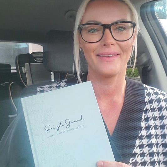 Woman holding a light blue book titled 'Surrogacy Journal' inside a car.